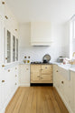 This small but functional kitchen features a white shaker design, complete with an L-shaped layout that includes a cooker and sink run topped with Carrara marble. It boasts a shaker-style canopy, glazed cupboards, drawers, and a white porcelain splashback. Additionally, there is a pantry and extra storage cupboards. The pale wooden plank flooring, aged brass handles, and cream gas stove add warmth to this bright and simple kitchen.