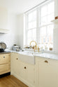 A close-up and angled view of a white linen shaker kitchen sink run featuring a double Belfast sink, an aged brass Ionian tap, and a Carrara marble worktop overlooking a glass window with a cityscape view.