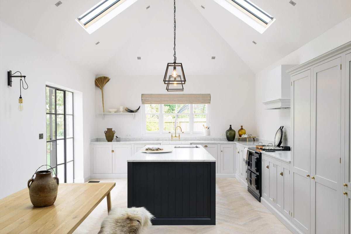 A spacious and artisan Shaker kitchen features an off-white L-shaped sink run with a double Belfast sink and an aged brass Ionian tap, a large pantry-blue kitchen island with a Carrara marble worktop, a shelf, a canopy, and a bank of cupboards. Cleverly decorated with steel-framed hanging lights, vintage ceramics, large glasses with narrow necks, and eclectic items serving as artistic displays, it creates a simple yet intriguing kitchen space.