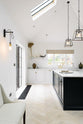 Nestled between an off-white L-shaped sink run with a double Belfast sink and an aged brass Ionian tap, and a pantry-blue kitchen island with a Carrara marble worktop, is a spacious and artisan Shaker kitchen. Cleverly decorated with steel-framed hanging lights, large glasses with narrow necks, vintage ceramics, and eclectic items serving as artistic displays, it creates a simple yet intriguing kitchen space.