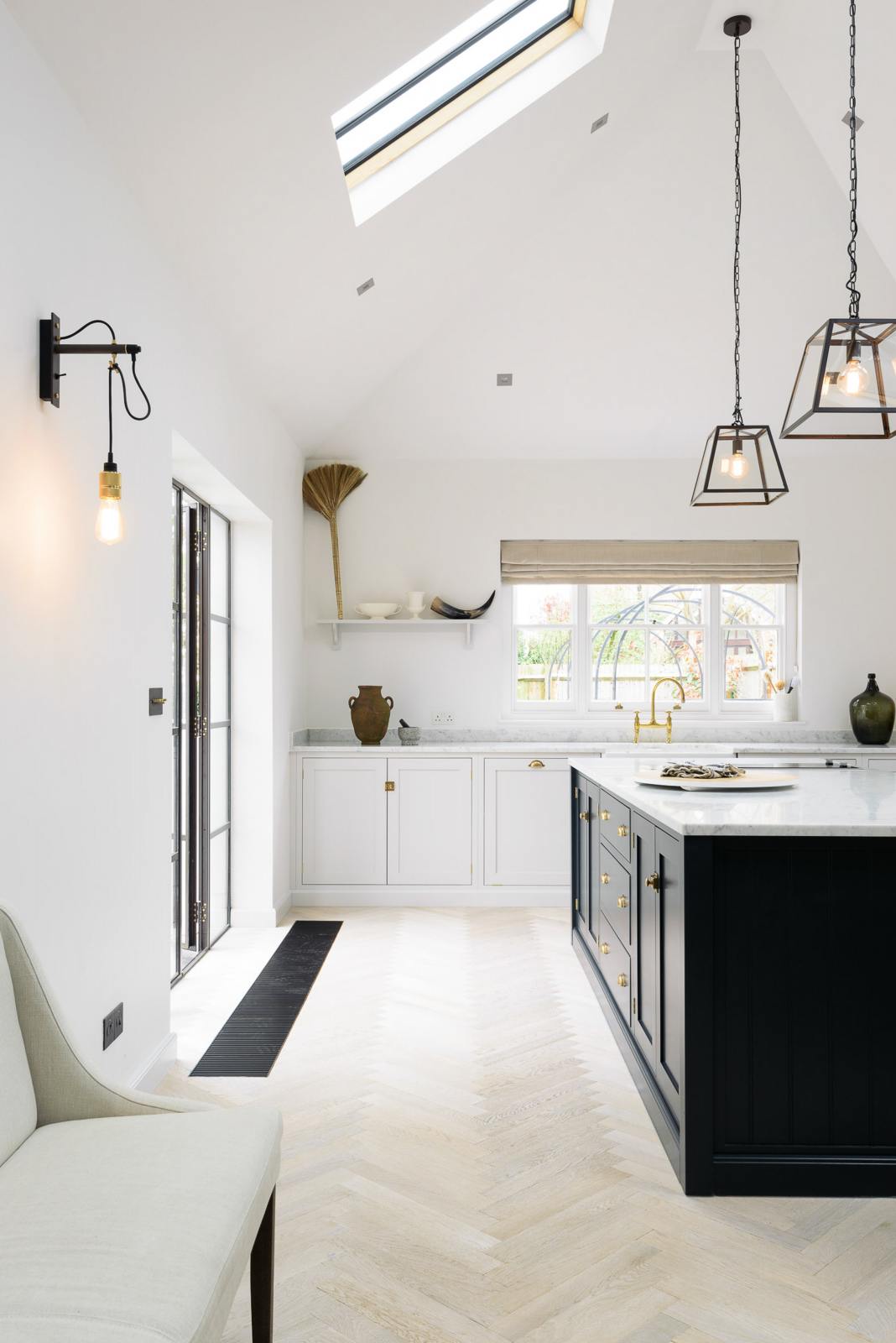 Nestled between an off-white L-shaped sink run with a double Belfast sink and an aged brass Ionian tap, and a pantry-blue kitchen island with a Carrara marble worktop, is a spacious and artisan Shaker kitchen. Cleverly decorated with steel-framed hanging lights, large glasses with narrow necks, vintage ceramics, and eclectic items serving as artistic displays, it creates a simple yet intriguing kitchen space.