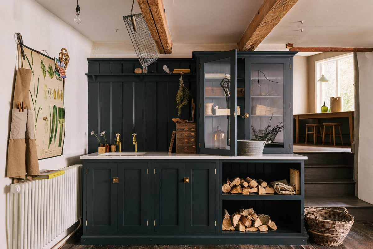 A sample pantry blue shaker kitchen’s sink run features a shelf, countertop glazed cupboards, and a mix of closed and open base cabinets. The Carrara marble worktop holds a copper single sink with aged brass Mayan taps. Wooden and metal accents—including firewood, a metal bin, and a wooden toolbox—add rustic charm.