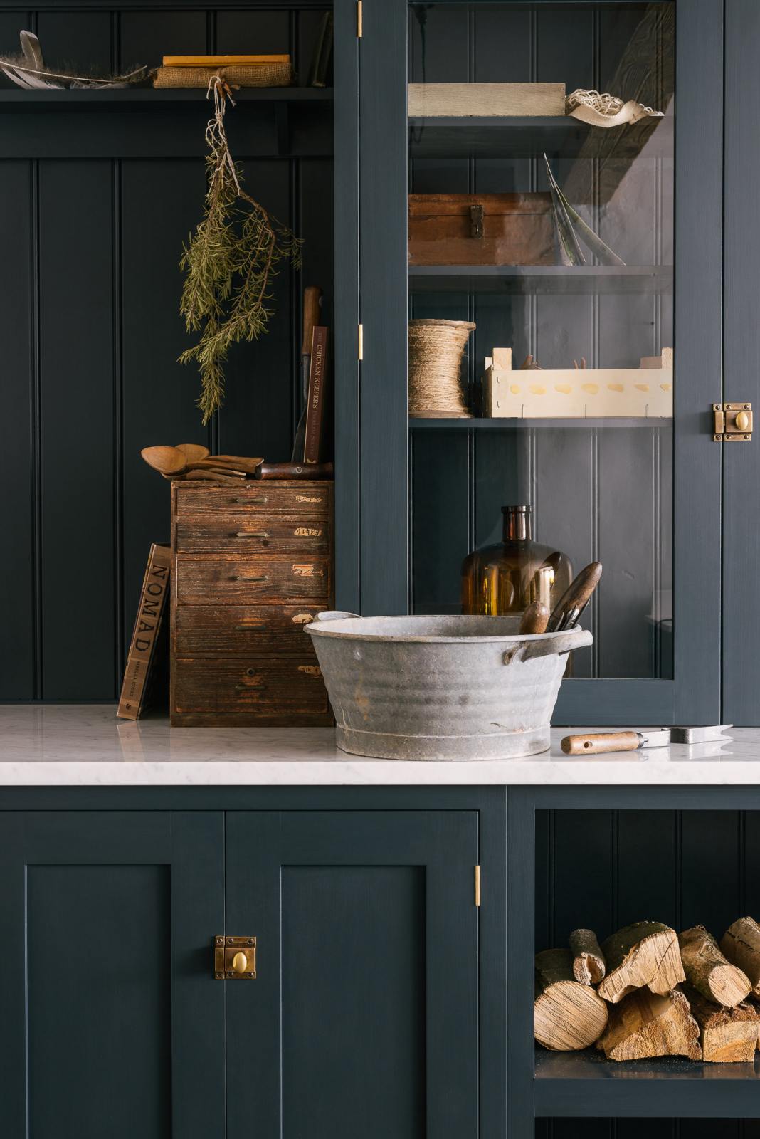 A closer look at the wooden and metal accents—including firewood, a metal bin, and a wooden toolbox of the pantry blue shaker kitchen’s sink run features a shelf, countertop glazed cupboards, a mix of closed and open base cabinets and a Carrara marble worktop.