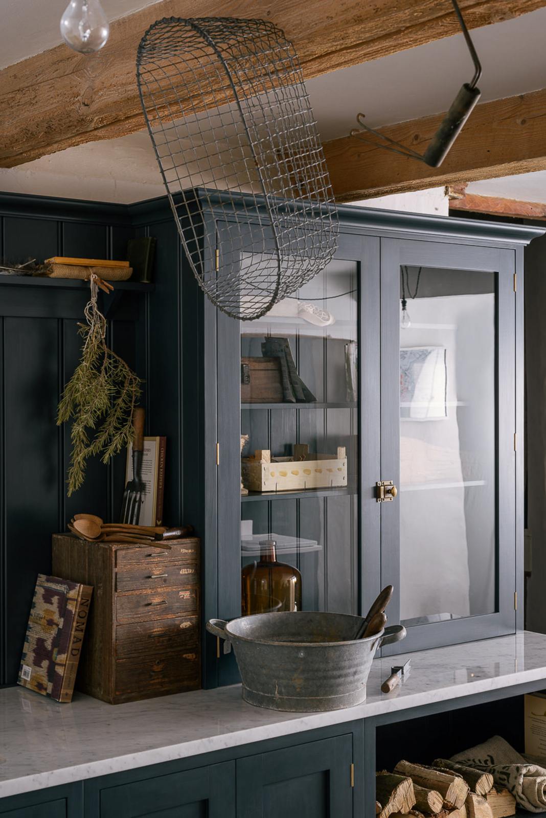A close and angled look at the wooden and metal accents—including firewood, a metal bin, and a wooden toolbox of the pantry blue shaker kitchen’s sink run features a shelf, countertop glazed cupboards, a mix of closed and open base cabinets and a Carrara marble worktop.