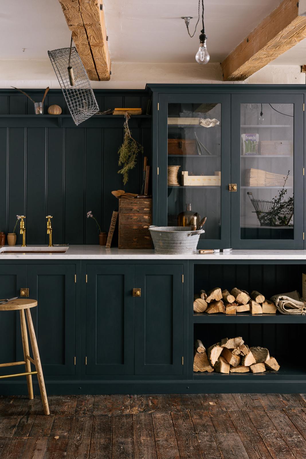 A closer look at the sample pantry blue shaker kitchen’s sink run features a shelf, countertop glazed cupboards, and a mix of closed and open base cabinets. The Carrara marble worktop holds a copper single sink with aged brass Mayan taps. Wooden and metal accents—including firewood, a metal bin, and a wooden toolbox—add rustic charm.