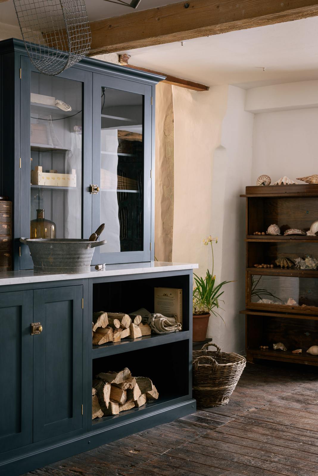 An angled look at the wooden and metal accents—including firewood, a metal bin, and a wooden toolbox of the pantry blue shaker kitchen’s sink run features a shelf, countertop glazed cupboards, a mix of closed and open base cabinets and a Carrara marble worktop.