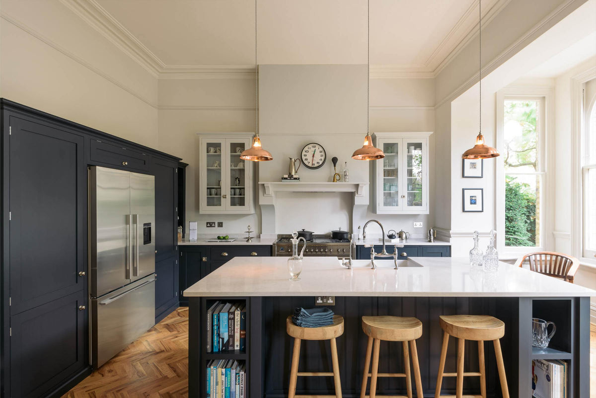 The pantry blue shaker kitchen features creamy Silestone worktops. A stainless steel fridge is built into the cabinetry. Off-white walls frame two pale wall cupboards flanking the chimney breast. The central island features a sink on one side and a breakfast bar with wooden stools on the other, beneath two hanging brass lights.