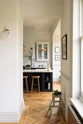 The pantry blue shaker kitchen features creamy Silestone worktops. Off-white walls frame two pale wall cupboards flanking the chimney breast. The central island features a sink on one side and a breakfast bar with wooden stools on the other. The kitchen and dining room have been combined into one large space, with a central column providing subtle separation.