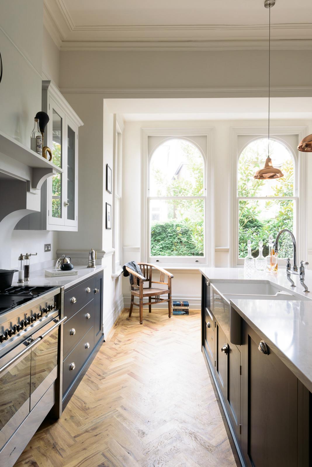 The pantry blue shaker kitchen features creamy Silestone worktops. Bright sunshine streams through the tall arched window, illuminating the central island, which houses a sink on one side and a breakfast bar on the other.