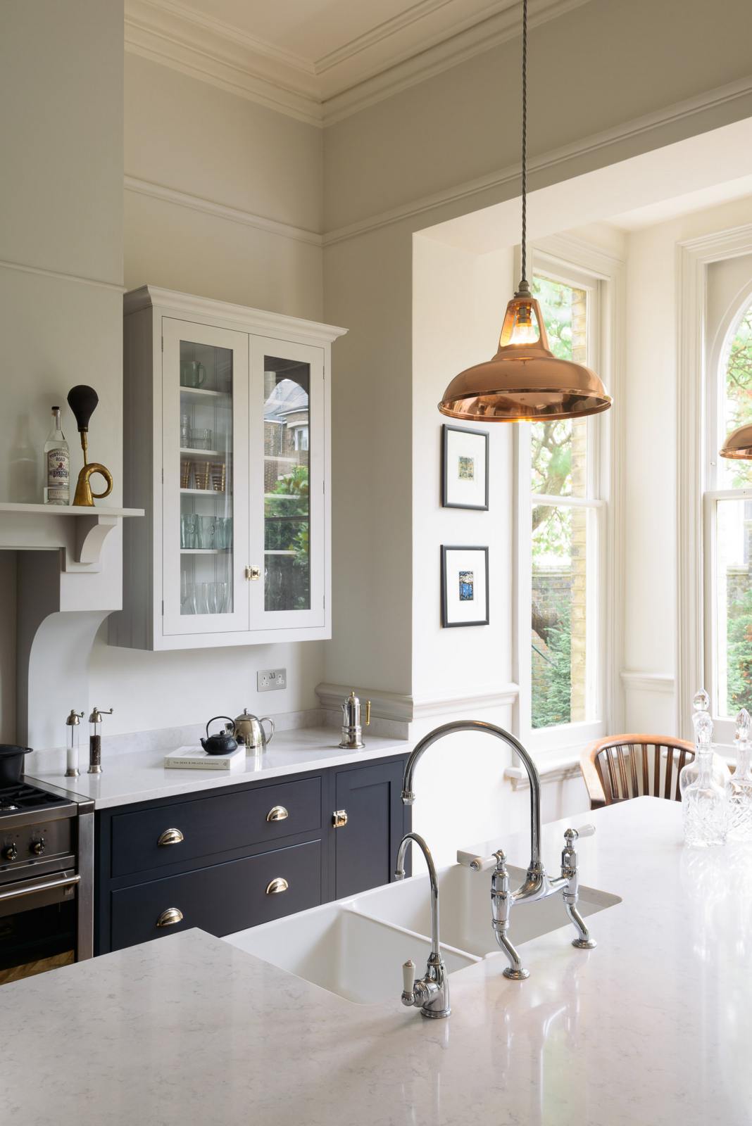 The pantry blue shaker kitchen features creamy Silestone worktops. The off-white walls feature a pale wall cupboard on the chimney breast. The central island houses a sink and a breakfast bar on the other side.