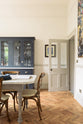 A door is open to welcome people, and a dark navy blue deVOL shaker cupboard stands next to it. In the foreground is a rustic table with vintage wooden chairs pulled up. The room features a herringbone-style wooden flooring and off-white walls. 