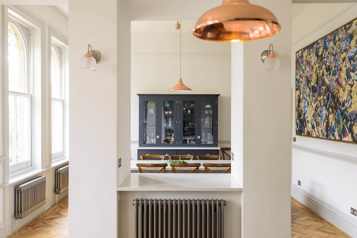 Two columns stand centre room, framing the dark navy blue cupboard. A rustic table with vintage wooden chairs sits before it, lit by a hanging light. The space features herringbone wooden floors and off-white walls.