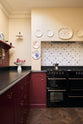 A quiet corner of a simple yet elegant Refectory red shaker kitchen features a long L-shaped sink and cooker run with a black granite worktop, a kitchen island, a tongue and groove splashback, a long shelf with brass rails, and a false chimney canopy over the cooker with blue delft splashback tiles. Beautifully decorated with artisan ceramics, antique champagne glasses, fresh produce, and wooden parquet flooring, creating a bold yet understated kitchen space.