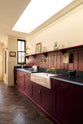An oblique-view of a simple yet elegant Refectory red shaker kitchen features a long L-shaped sink and cooker run with a black granite worktop, a tongue and groove splashback, and a long shelf with brass rails. Beautifully decorated with artisan ceramics, antique champagne glasses, an oil painting, and fresh fruits, creating a bold yet understated kitchen space.