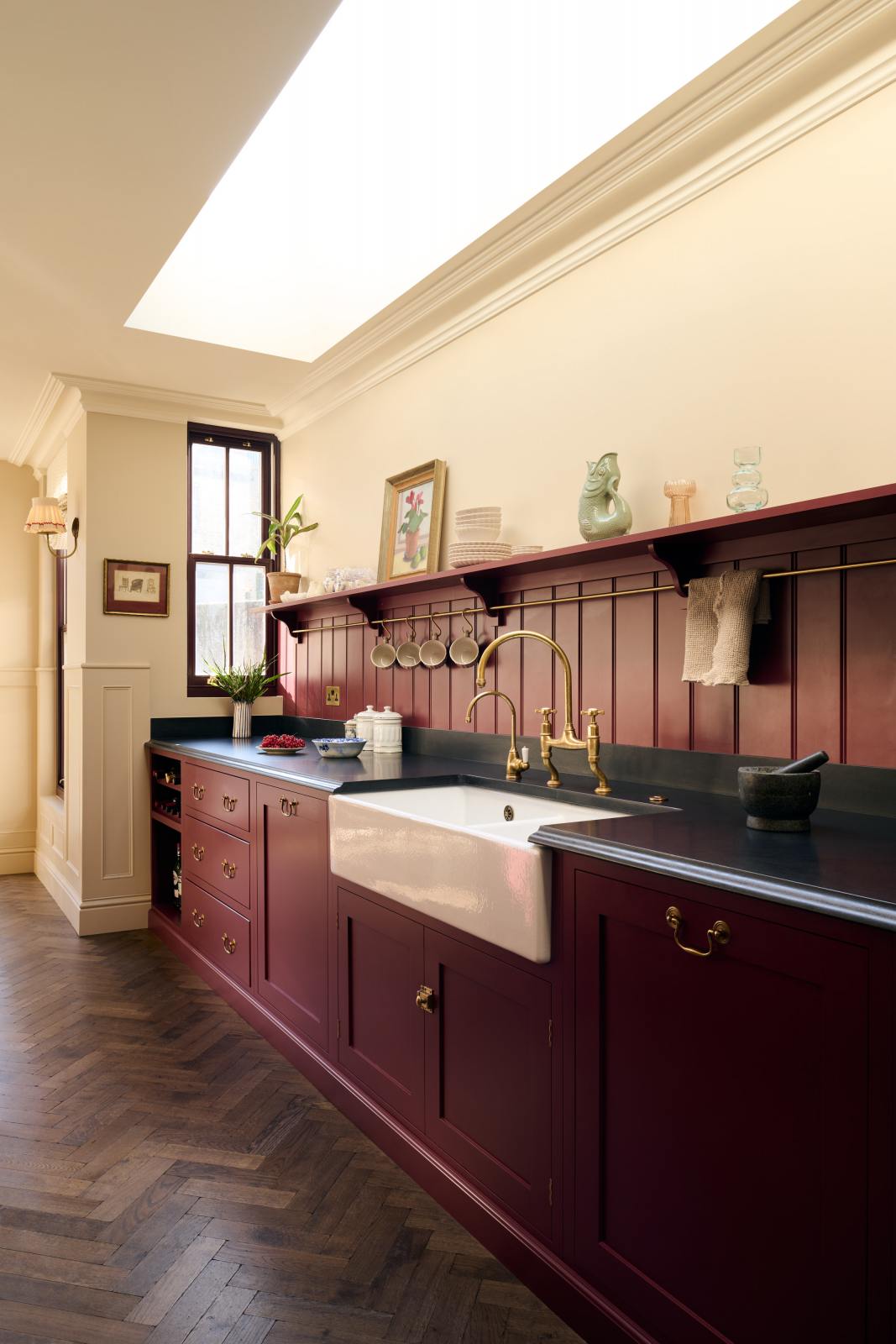 An oblique-view of a simple yet elegant Refectory red shaker kitchen features a long L-shaped sink and cooker run with a black granite worktop, a tongue and groove splashback, and a long shelf with brass rails. Beautifully decorated with artisan ceramics, antique champagne glasses, an oil painting, and fresh fruits, creating a bold yet understated kitchen space.
