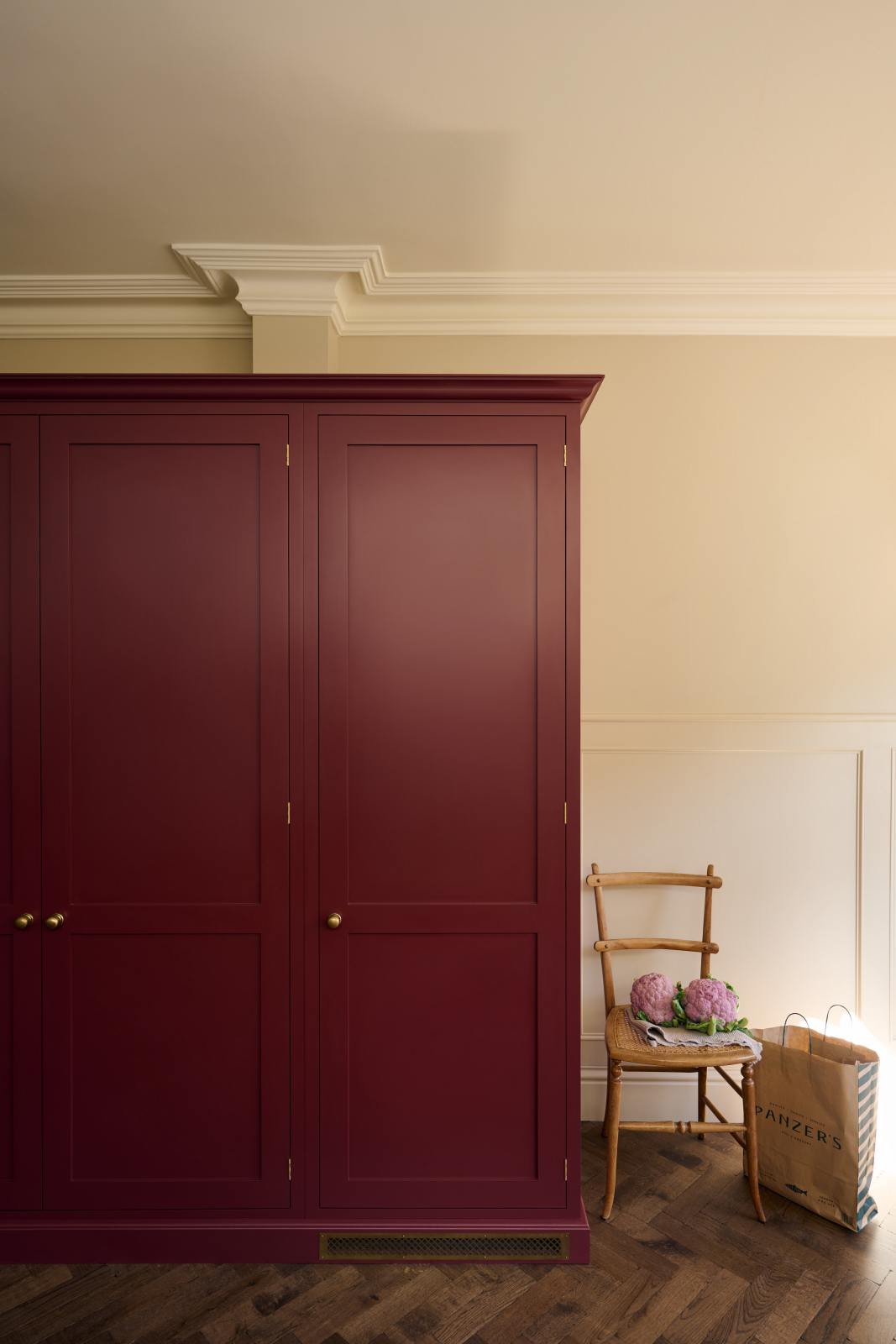 A quiet corner of a simple yet elegant Refectory red shaker kitchen features a bank of pantry cupboards. Beautifully decorated with a wooden chair, red cauliflower, a brown shopping bag, and wooden parquet flooring, creating a bold yet understated kitchen space.