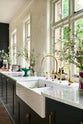 An oblique view of the double Belfast sink with an aged brass Ionian tap in the Bakehouse Green shaker kitchen’s sink run, featuring Carrara marble worktop. Beautifully decorated with tall, white wildflowers in found glass jars, which add colour to the off-white, delicately patterned wallpaper.