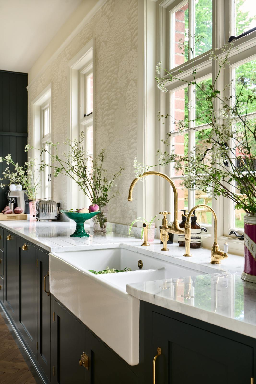 An oblique view of the double Belfast sink with an aged brass Ionian tap in the Bakehouse Green shaker kitchen’s sink run, featuring Carrara marble worktop. Beautifully decorated with tall, white wildflowers in found glass jars, which add colour to the off-white, delicately patterned wallpaper.