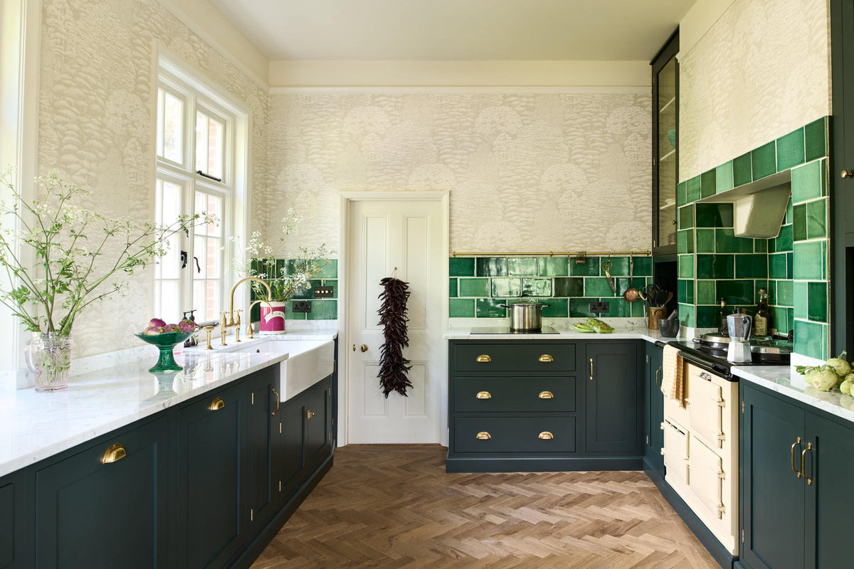 This spacious Bakehouse Green shaker kitchen features a sink run and a concave cooker run, complemented by a Carrara marble worktop. It features a solid oak parquet floor, glazed cabinet, an aged brass hanging rail and Emerald Green London splashback tiles, which add colour to the off-white, delicately patterned wallpaper.