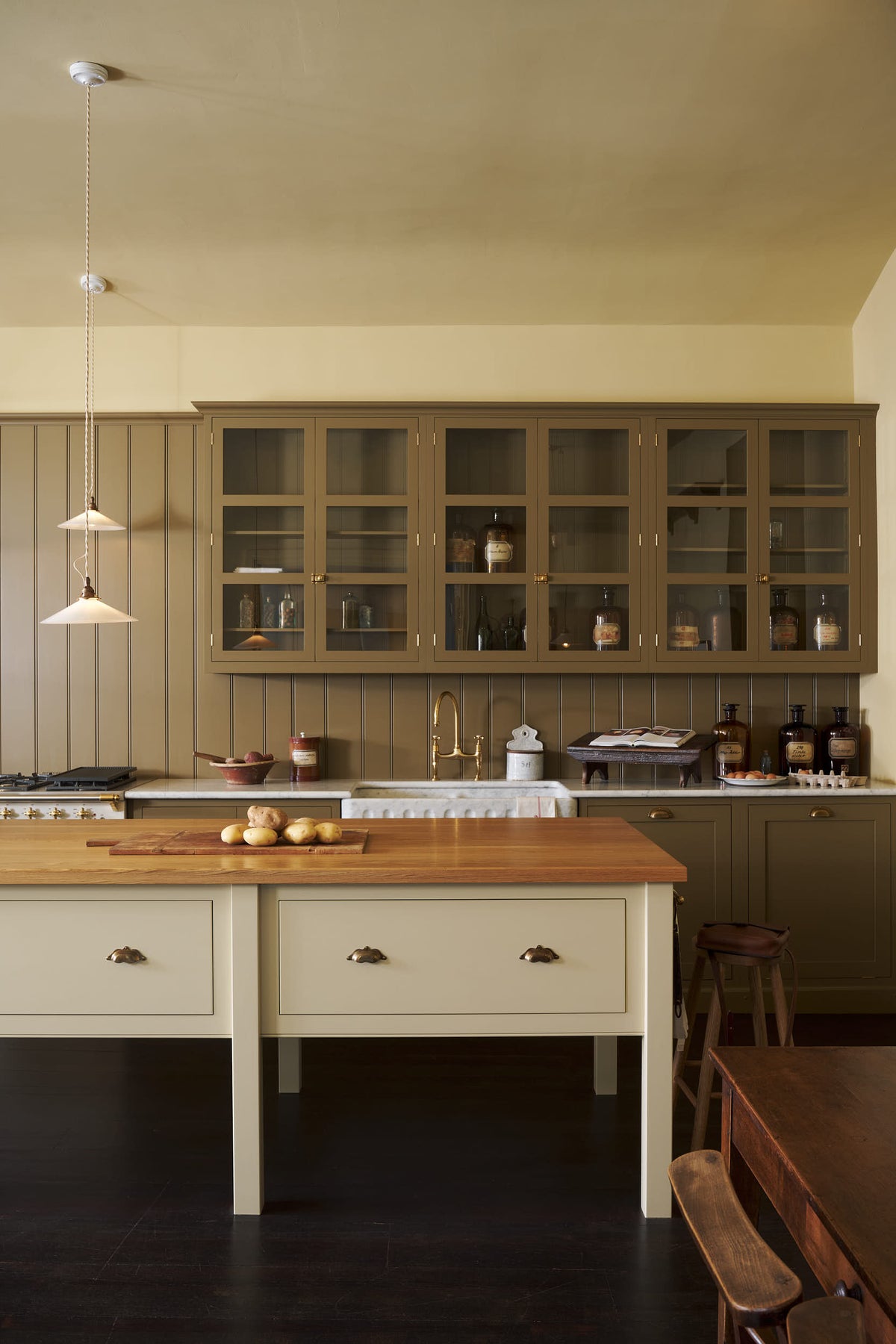 A quiet corner of a simple Quaker cream and Folgate green Shaker kitchen features a cooker and sink run with Carrara marble worktop, glazed wall cupboards, and a bespoke prep table with a classic oak worktop. Lovingly adorned with milk pendant lights, artisan ceramics, fresh potatoes, and antique furniture, it creates a warm and inviting atmosphere.