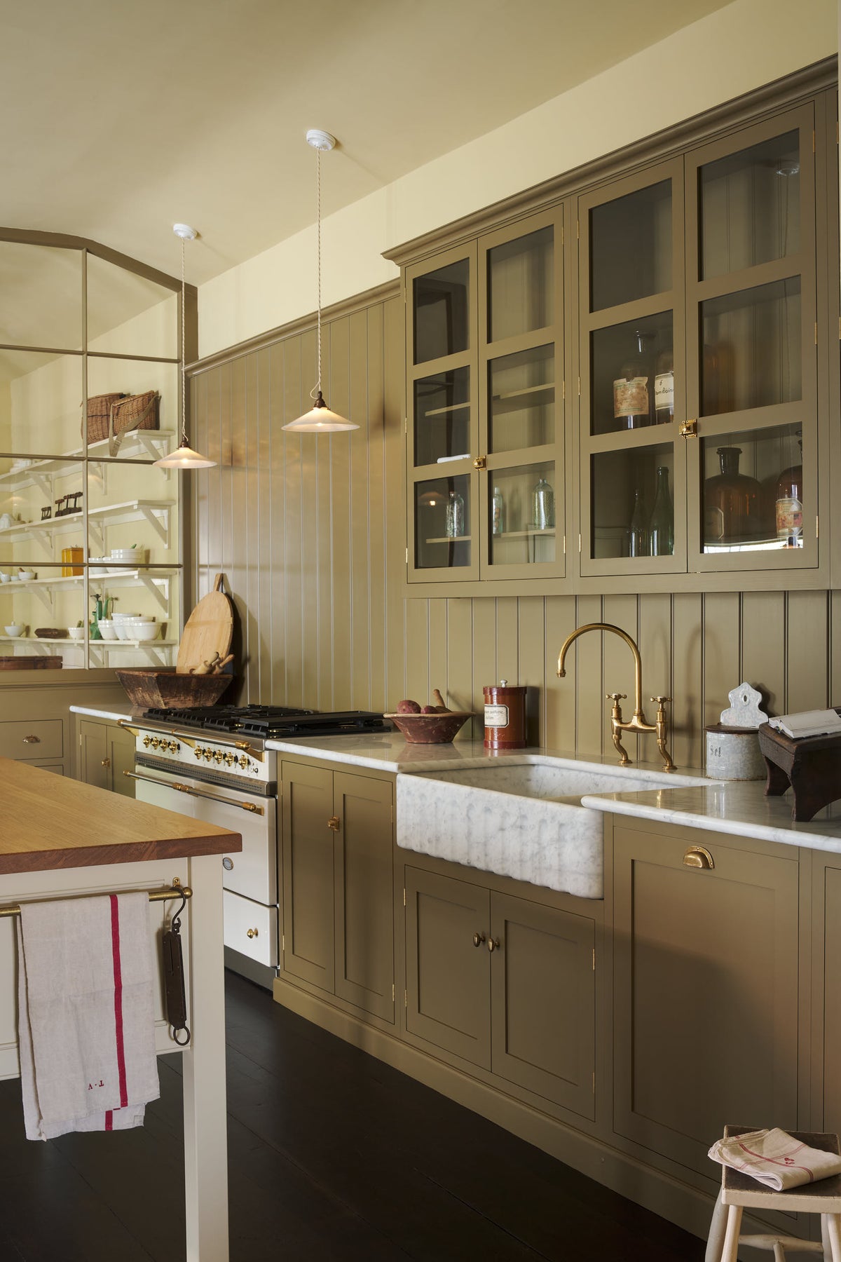 An oblique view of a simple Quaker cream and Folgate green Shaker kitchen features a cooker and sink run with Carrara marble worktop, glazed wall cupboards, and a bespoke prep table with a classic oak worktop. Lovingly adorned with milk pendant lights, artisan ceramics, and antique furniture, it creates a warm and inviting atmosphere.