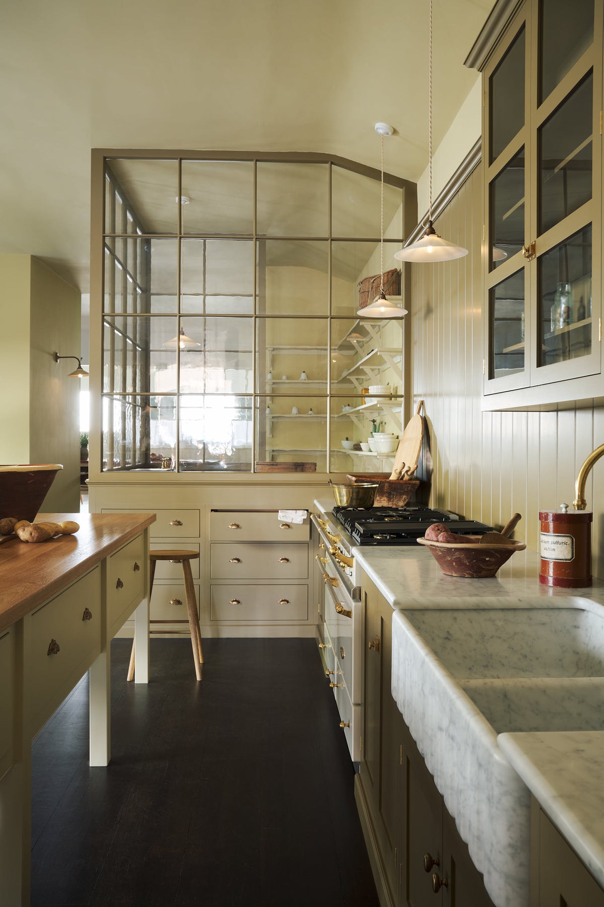 A simple Quaker cream and Folgate green Shaker kitchen features a cooker and sink run with Carrara marble worktop, glazed wall cupboards, and a bespoke prep table with a classic oak worktop. Lovingly adorned with milk pendant lights, artisan ceramics, antiques, and a Helen stool, it creates a warm and inviting atmosphere.