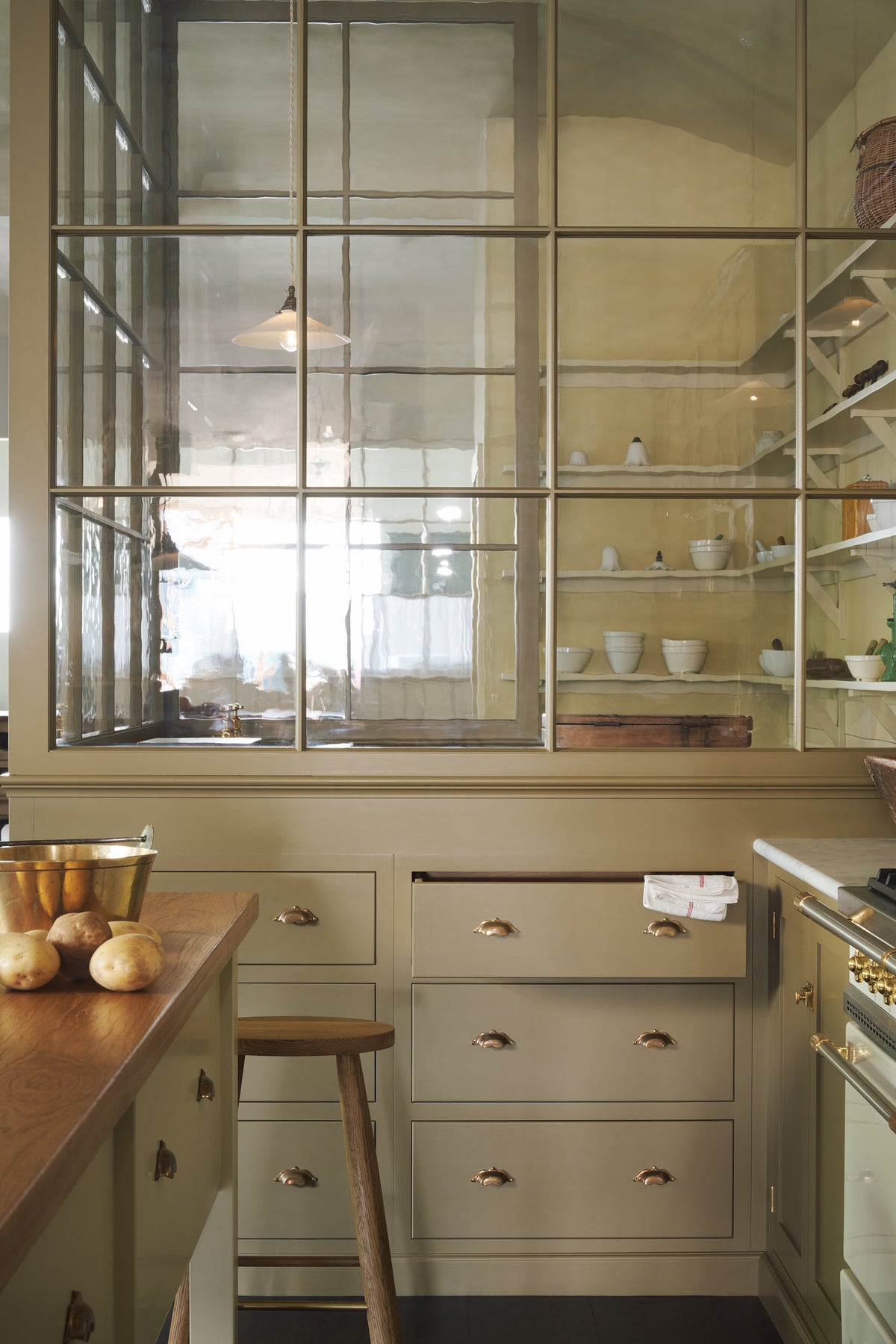 A closer look at a simple Quaker cream and Folgate green Shaker kitchen features a cooker and sink run with Carrara marble worktop, pan drawers, and a bespoke prep table with a classic oak worktop. Lovingly adorned with a tea towel, potatoes, antiques, and a Helen stool, it creates a warm and inviting atmosphere.
