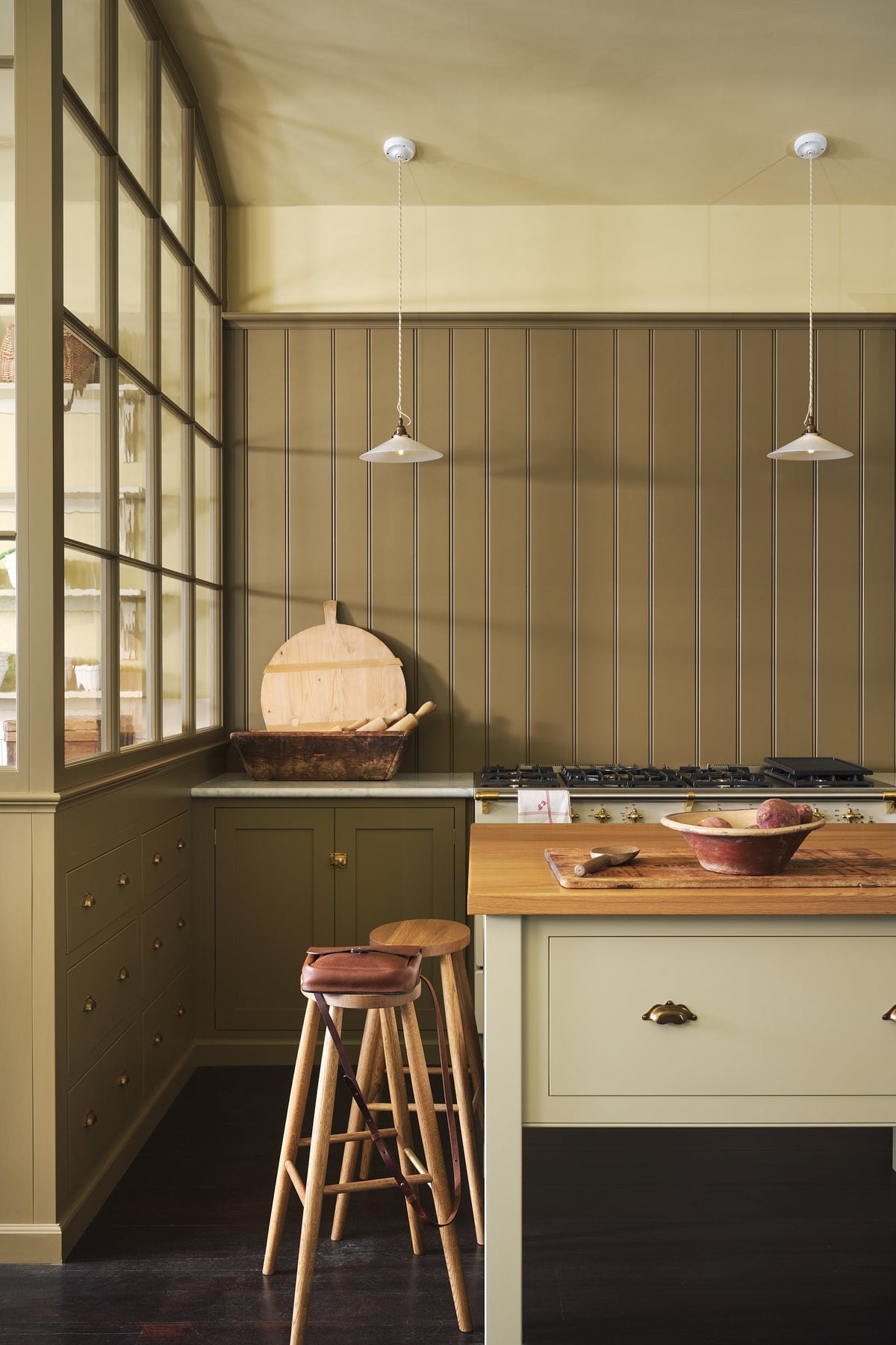 A quiet corner of a simple Quaker cream and Folgate green Shaker kitchen features a cooker and sink run with Carrara marble worktop, and a bespoke prep table with a classic oak worktop. Lovingly adorned with milk pendant lights, artisan ceramics, fresh potatoes, Helen stools, and antiques, it creates a warm and inviting atmosphere.