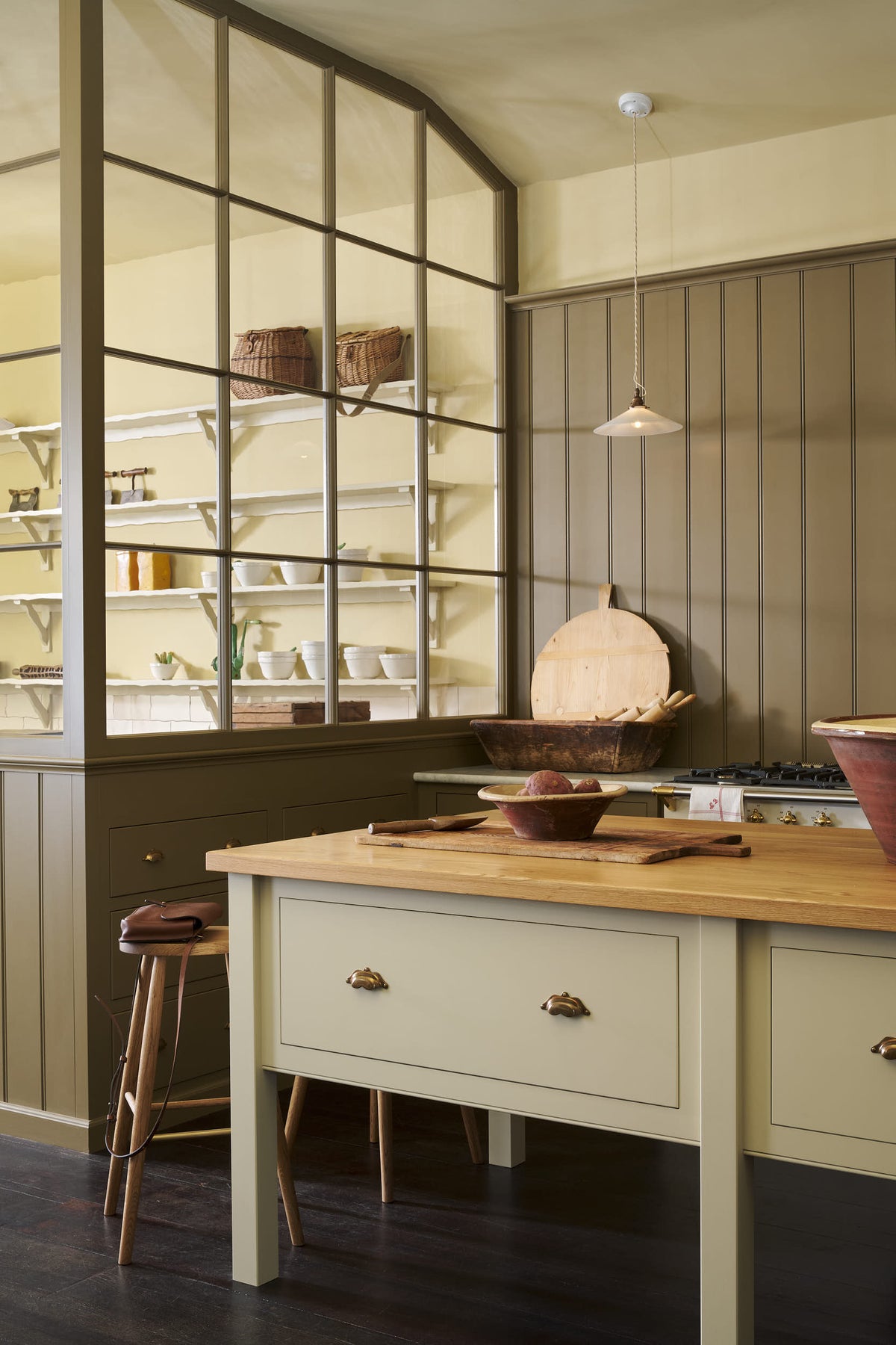An oblique view of a quiet corner of a simple Quaker cream and Folgate green Shaker kitchen features a cooker and sink run with Carrara marble worktop, and a bespoke prep table with a classic oak worktop. Lovingly adorned with milk pendant lights, artisan ceramics, fresh potatoes, Helen stools, and antiques, it creates a warm and inviting atmosphere.