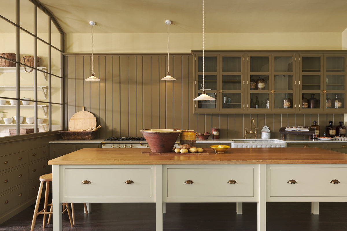 A simple Quaker cream and Folgate green Shaker kitchen features a cooker and sink run with Carrara marble worktop, glazed wall cupboards, and a bespoke prep table with a classic oak worktop. Lovingly adorned with milk pendant lights, artisan ceramics, antiques, and Helen stools, it creates a warm and inviting atmosphere.