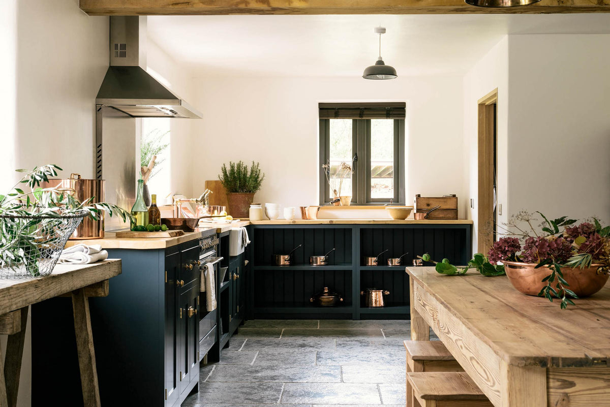 A charming cottage shaker kitchen in Pantry Blue features a cooker run, open base cabinets, and a solid oak worktop that complements the wooden ceiling beams and the vintage dining table with stools.
