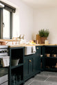 A closer look at the charming Pantry Blue cottage Shaker kitchen reveals a cooker run, open base cabinets, and a solid oak worktop that complements the wooden ceiling beams and vintage dining table with stools. The open cabinets showcase rose-gold cookware, reflecting the warmth of the wood while offering easy, functional access.
