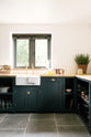 A closer look at the charming Pantry Blue Shaker kitchen reveals a cooker run, open base cabinets, and a solid oak worktop that echoes the wooden ceiling beams and vintage dining table with stools. The open cabinets display rose-gold cookware, adding warmth that complements the wooden accents.