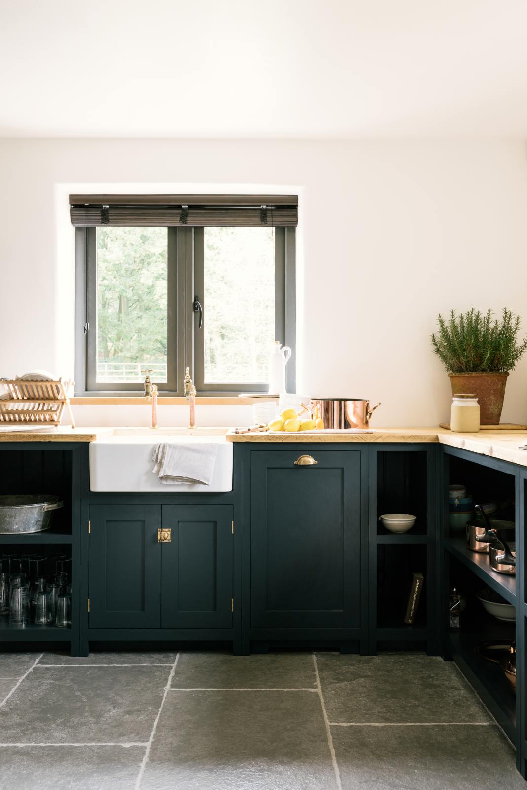A closer look at the charming Pantry Blue Shaker kitchen reveals a cooker run, open base cabinets, and a solid oak worktop that echoes the wooden ceiling beams and vintage dining table with stools. The open cabinets display rose-gold cookware, adding warmth that complements the wooden accents.