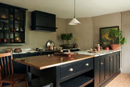 An oblique view of an understated painter’s black shaker kitchen featuring an aged copper sink, aged brass taps, and a San Simone quartzite worktop—its pale white base accented with bold dark grey veins and subtle gold flecks. Grey walls frame the space, with a cheese plant and vintage oil painting adding character in the background.