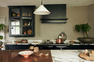 A straight view of an understated painter’s black shaker kitchen featuring an aged copper sink, aged brass taps, and a San Simone quartzite worktop—its pale white base accented with bold dark grey veins and subtle gold flecks. Grey walls frame the space, with a cheese plant and vintage oil painting adding character in the background.