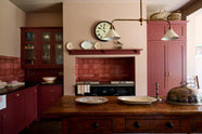 A humble Refectory red shaker kitchen featuring a sink run, glazed cupboards, a full-height pantry cupboard, and a large gas cooker in a chimney breast. The kitchen’s striking visuals are the Red Lace Market tiles used as a splashback in the chimney breast and along the sink run, enhancing the room's warmth. The space is decorated with vintage furniture, particularly an old, solid oak prep table.