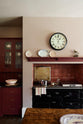 A humble Refectory red shaker kitchen featuring a sink run, glazed cupboards, a full-height pantry cupboard, and a large gas cooker in a chimney breast. The kitchen’s striking visuals are the Red Lace Market tiles used as a splashback in the chimney breast and along the sink run, enhancing the room's warmth. The space is decorated with vintage furniture, particularly an old, solid oak prep table.