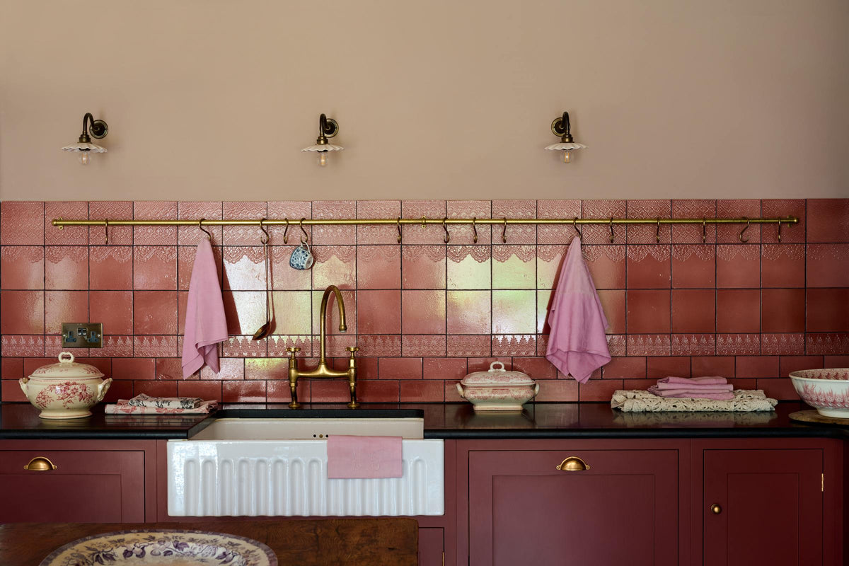 A closer look at a modest Refectory red shaker kitchen, featuring a sink with a granite worktop, a single fluted Belfast sink with an aged brass Ionian tap, and Red Lace Market splashback tiles complemented by aged brass hanging rails for holding cooking utensils and tea towels.
