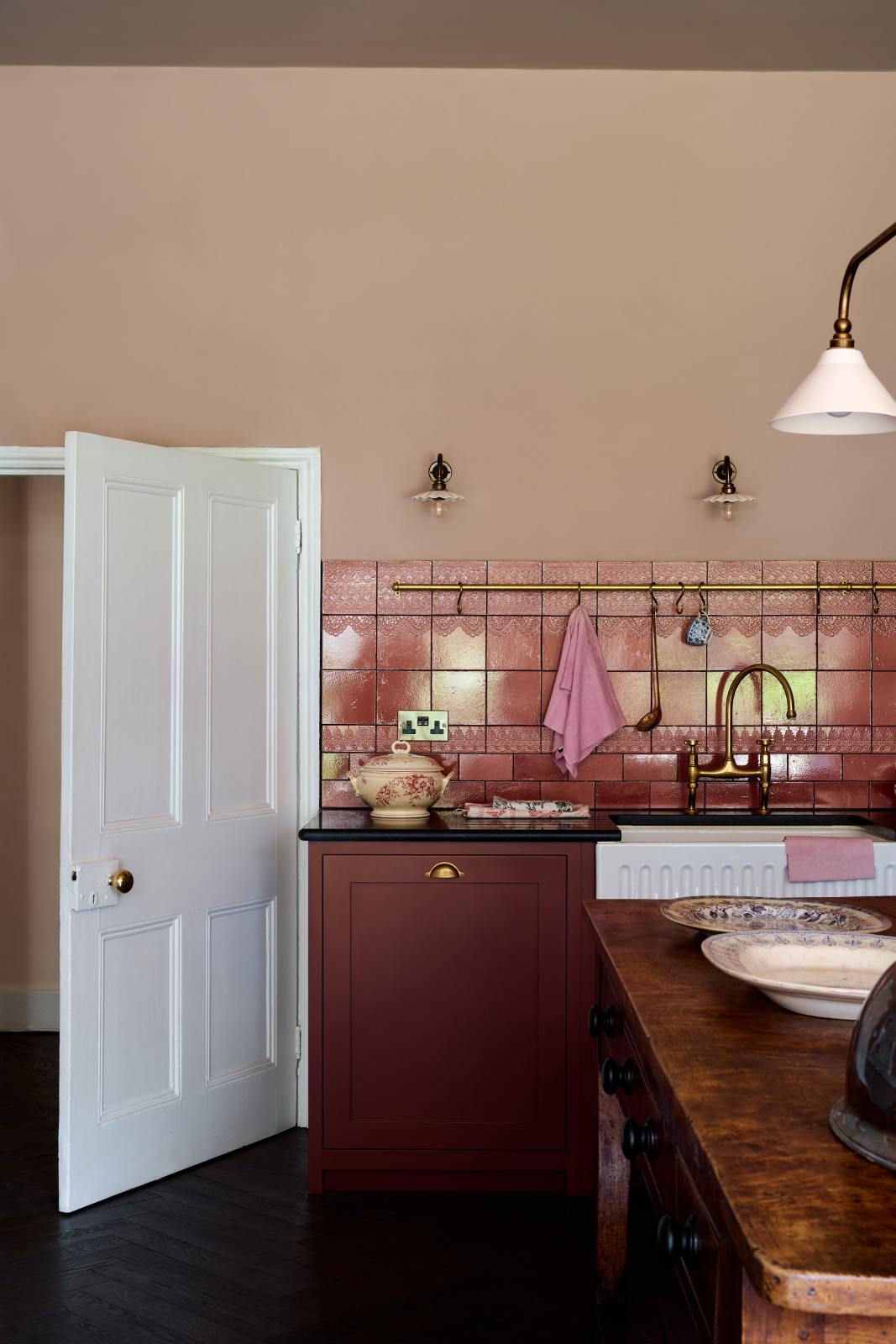 A corner of a modest Refectory Red shaker kitchen, featuring a sink with a granite worktop, a single fluted Belfast sink with an aged brass Ionian tap, and Red Lace Market splashback tiles complemented by aged brass hanging rails for holding cooking utensils and tea towels.