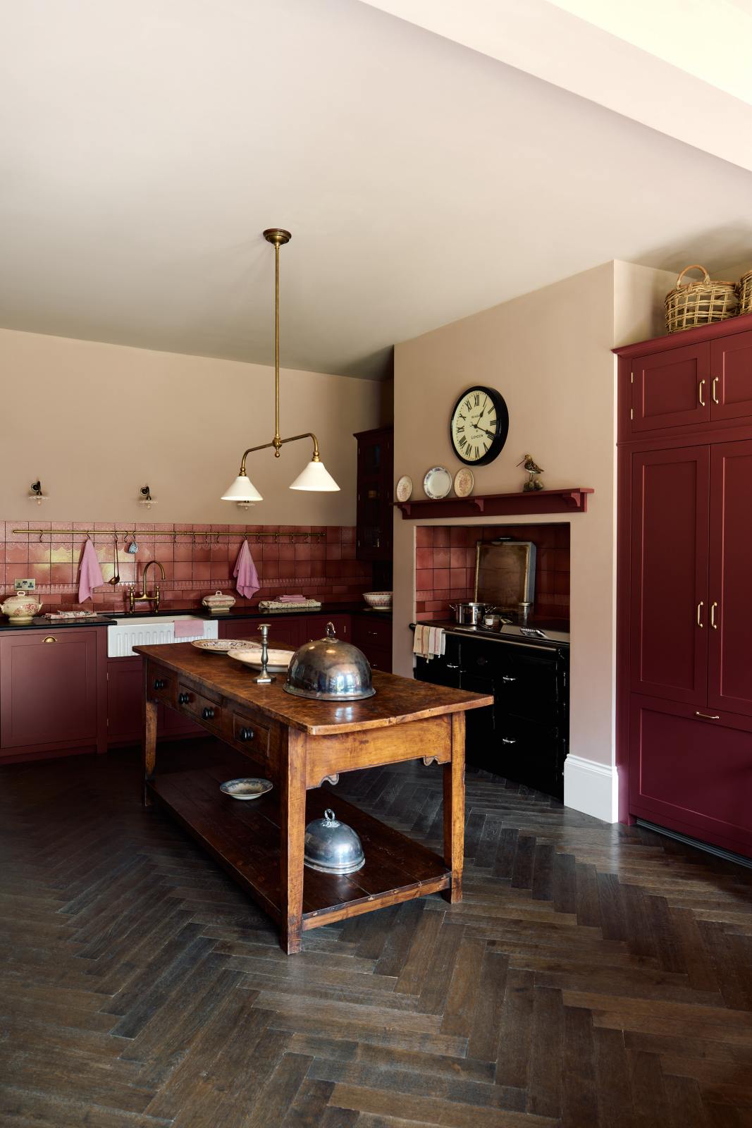 An angled view of a humble Refectory red shaker kitchen featuring a sink run, glazed cupboards,  a full-height pantry cupboard, and a large gas cooker in a chimney breast. The kitchen’s striking visuals are the Red Lace Market tiles used as a splashback in the chimney breast and along the sink run, enhancing the room's warmth. The space is decorated with vintage furniture, particularly an old, solid oak prep table and beautiful rich oak parquet floors.