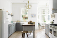 A light and bright Shaker kitchen features an L-shaped sink run with a double sink, an aged brass Ionian tap, a cooker run with a canopy, Silestone worktop, and a Danmask prep table with a solid oak worktop. Beautifully decorated with soft still-life paintings, a white refrigerator, a modern brass chandelier, vintage furniture, and an adorable set of soft toys sitting at the edge of a breakfast nook, it creates a wonderfully warm and contemporary space.