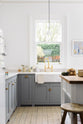 A light and bright Shaker kitchen features an L-shaped sink run with a double sink, an aged brass Ionian tap, Silestone worktop, and a Danmask prep table with a solid oak worktop. Beautifully decorated with soft still-life paintings, a white refrigerator, a modern brass chandelier, and vintage furniture, it creates a wonderfully warm and contemporary space.