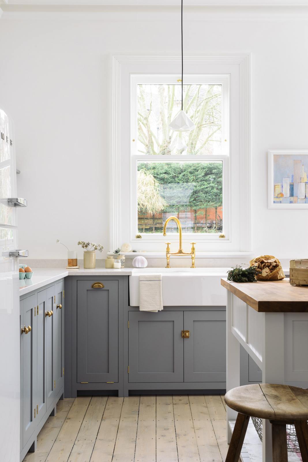 A light and bright Shaker kitchen features an L-shaped sink run with a double sink, an aged brass Ionian tap, Silestone worktop, and a Danmask prep table with a solid oak worktop. Beautifully decorated with soft still-life paintings, a white refrigerator, a modern brass chandelier, and vintage furniture, it creates a wonderfully warm and contemporary space.