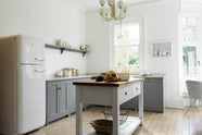 A light and bright Shaker kitchen features an L-shaped sink run with a double sink, an aged brass Ionian tap, Silestone worktop, and a Danmask prep table with a solid oak worktop. Beautifully decorated with soft still-life paintings, a white refrigerator, a modern brass chandelier, vintage furniture, and a breakfast nook, it creates a wonderfully warm and contemporary space.