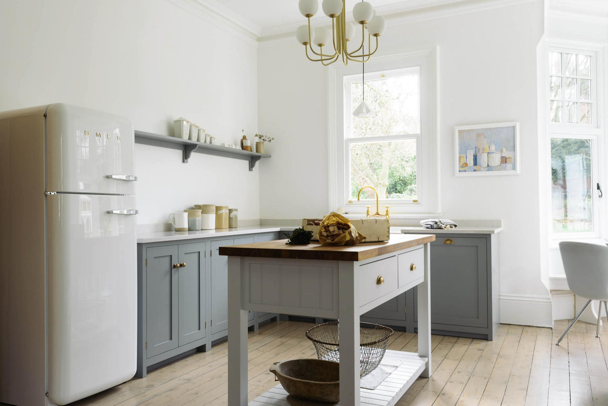 A light and bright Shaker kitchen features an L-shaped sink run with a double sink, an aged brass Ionian tap, Silestone worktop, and a Danmask prep table with a solid oak worktop. Beautifully decorated with soft still-life paintings, a white refrigerator, a modern brass chandelier, vintage furniture, and a breakfast nook, it creates a wonderfully warm and contemporary space.