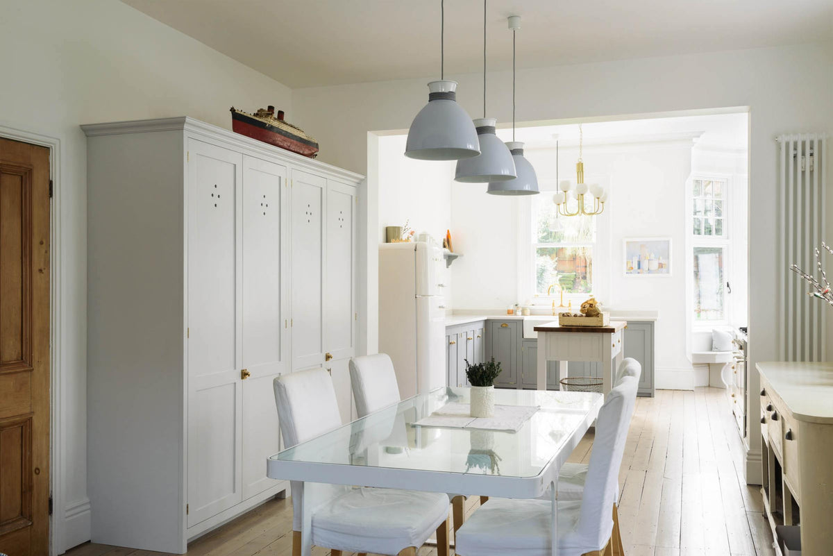 An angled view of a light dining room features double pantry cupboards, and through the archway is a bright Shaker kitchen with an L-shaped sink run that includes a double sink, an aged brass Ionian tap, a Silestone worktop, and a Danmask prep table with a solid oak worktop. Beautifully decorated with soft still-life paintings, a white refrigerator, a modern brass chandelier, and vintage furniture, it creates a wonderfully warm and contemporary space.