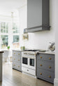 An oblique view of a light and bright Shaker kitchen features a cooker run with a canopy, Silestone worktop, and a Danmask prep table with a solid oak worktop. Beautifully decorated with soft still-life paintings, vintage furniture, and a breakfast nook, it creates a wonderfully warm and contemporary space.