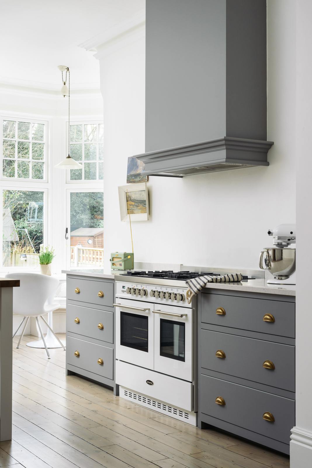 An oblique view of a light and bright Shaker kitchen features a cooker run with a canopy, Silestone worktop, and a Danmask prep table with a solid oak worktop. Beautifully decorated with soft still-life paintings, vintage furniture, and a breakfast nook, it creates a wonderfully warm and contemporary space.