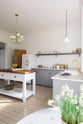 An angled view of a light and bright Shaker kitchen features an L-shaped sink run with a double sink, an aged brass Ionian tap, Silestone worktop, a long shelf and a Danmask prep table with a solid oak worktop. Beautifully decorated with a white refrigerator, a modern brass chandelier, and vintage furniture, it creates a wonderfully warm and contemporary space.